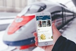 A person is holding a smartphone displaying the LiveJournal app, with a sleek, modern high-speed train positioned in the background at the station. The phone screen shows various posts including text and images.