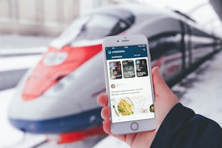 A person is holding a smartphone displaying the LiveJournal app, with a sleek, modern high-speed train positioned in the background at the station. The phone screen shows various posts including text and images.