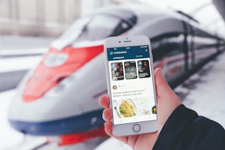 A person is holding a smartphone displaying the LiveJournal app, with a sleek, modern high-speed train positioned in the background at the station. The phone screen shows various posts including text and images.