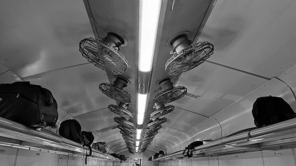 The image depicts a train car interior with white walls and ceiling, illuminated by multiple overhead fans that are turned on. The perspective is from the floor, looking up towards the ceiling, providing an upward view of the space. On the lower level, there are several rows of seats lined up against the wall, each equipped with a black suitcase resting on it.