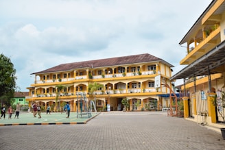 Three-story yellow building with balconies and a basketball hoop in the courtyard, surrounded by children playing. The sky is partly cloudy.