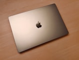 A closed silver laptop with a distinctive logo in the center is resting on a wooden surface. The lighting highlights the sleek and modern design of the device.