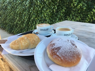 A wooden table set in an outdoor space features two cups of coffee with frothy tops and branding from Dinzler. There is a croissant and a pastry dusted with powdered sugar on separate plates, both laid on napkins. The background is dominated by a lush green hedge.