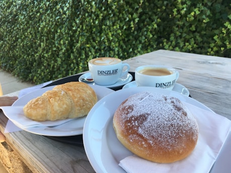 A wooden table set in an outdoor space features two cups of coffee with frothy tops and branding from Dinzler. There is a croissant and a pastry dusted with powdered sugar on separate plates, both laid on napkins. The background is dominated by a lush green hedge.