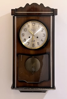 A vintage wall clock with a wooden frame and a round silver clock face. The clock has Arabic numerals and gold-colored hands. Below the clock face is a decorative section with a circular design and a glass cover.