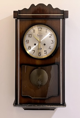 A vintage wall clock with a wooden frame and a round silver clock face. The clock has Arabic numerals and gold-colored hands. Below the clock face is a decorative section with a circular design and a glass cover.