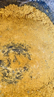 A close-up view of a coarse, yellowish-brown powder spread across a dark surface. The texture appears rough, with uneven clumps and scattered fine particles of the same material.