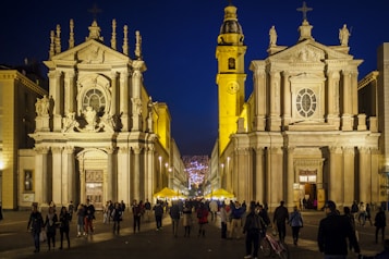 A nighttime cityscape featuring a grand Baroque-style church with intricate architectural details. The illuminated facade is complemented by an adjacent bell tower, both exuding a warm glow. People leisurely stroll in the foreground, adding life to the scene, while festive lights in the distance suggest a lively atmosphere.