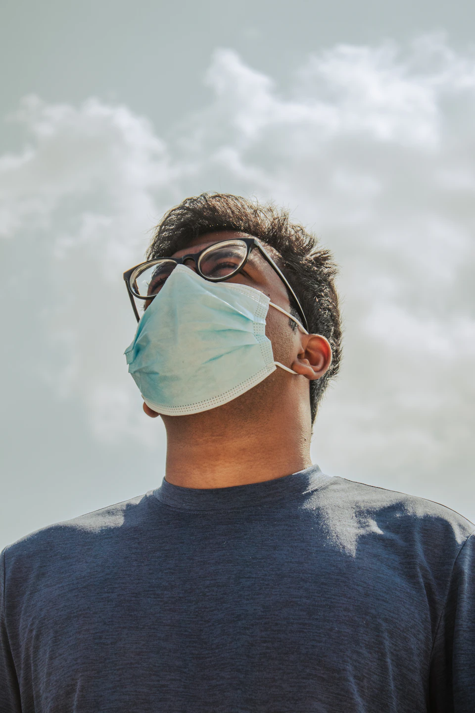 Man wearing glasses and blue surgical mask stands against cloudy sky with masked face