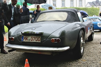 A gray vintage convertible car with a German license plate is parked on a gravel driveway. Several people are gathered around, with some holding umbrellas, indicating it might be raining or has recently rained. Another car can be seen in the background, and there are some orange traffic cones nearby.