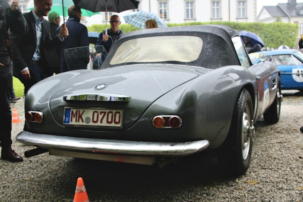 A gray vintage convertible car with a German license plate is parked on a gravel driveway. Several people are gathered around, with some holding umbrellas, indicating it might be raining or has recently rained. Another car can be seen in the background, and there are some orange traffic cones nearby.