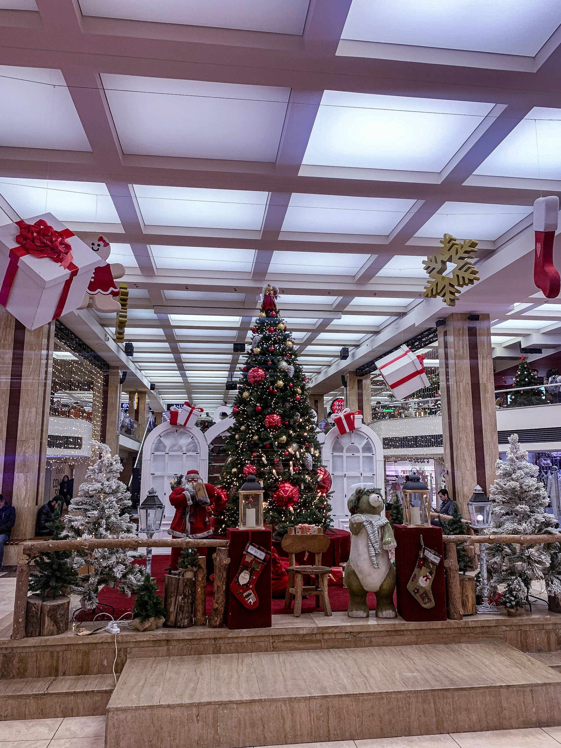 A festive indoor scene featuring an elaborately decorated Christmas tree with red and gold ornaments and a star on top. Surrounding the tree are white artificial trees with fake snow, several large gift boxes hanging from the ceiling, a Santa Claus figure, and a plush bear dressed in winter attire. The setting appears to be inside a shopping mall, with additional Christmas decorations like stockings and lanterns placed around.
