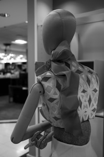 A monochrome image of a mannequin dressed in an intricately patterned top with a large bow at the back. The wooden mannequin is positioned in a retail environment with blurred store lights and shelves in the background.