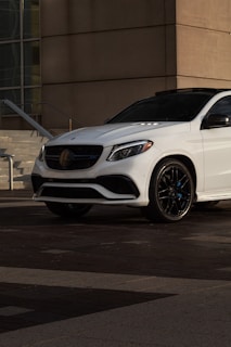 A white luxury SUV is parked on a modern urban street in front of a building with geometric architectural elements. The car displays a sleek design with tinted windows and shiny black wheels. The lighting creates a shadow on the ground, suggesting either early morning or late afternoon.