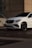 A white luxury SUV is parked on a modern urban street in front of a building with geometric architectural elements. The car displays a sleek design with tinted windows and shiny black wheels. The lighting creates a shadow on the ground, suggesting either early morning or late afternoon.
