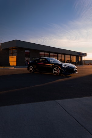 A sleek black car is parked in front of a modern building with large windows reflecting the sunset. The sky is a soft gradient of blue and orange, creating a serene atmosphere. The building has a flat roof and brick exterior, with the surroundings mostly deserted.