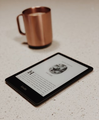 A Kindle e-reader displays a page from a book, laid on a light-colored speckled countertop. In the background, a copper-colored mug with a handle is slightly out of focus.