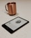 A Kindle e-reader displays a page from a book, laid on a light-colored speckled countertop. In the background, a copper-colored mug with a handle is slightly out of focus.