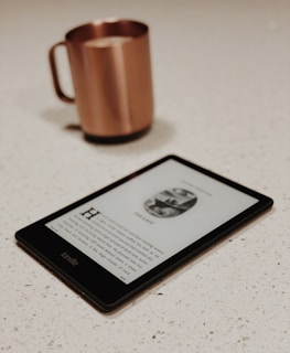 A Kindle e-reader displays a page from a book, laid on a light-colored speckled countertop. In the background, a copper-colored mug with a handle is slightly out of focus.