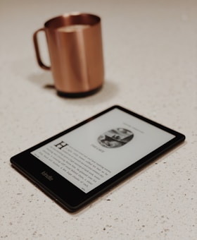 A Kindle e-reader displays a page from a book, laid on a light-colored speckled countertop. In the background, a copper-colored mug with a handle is slightly out of focus.