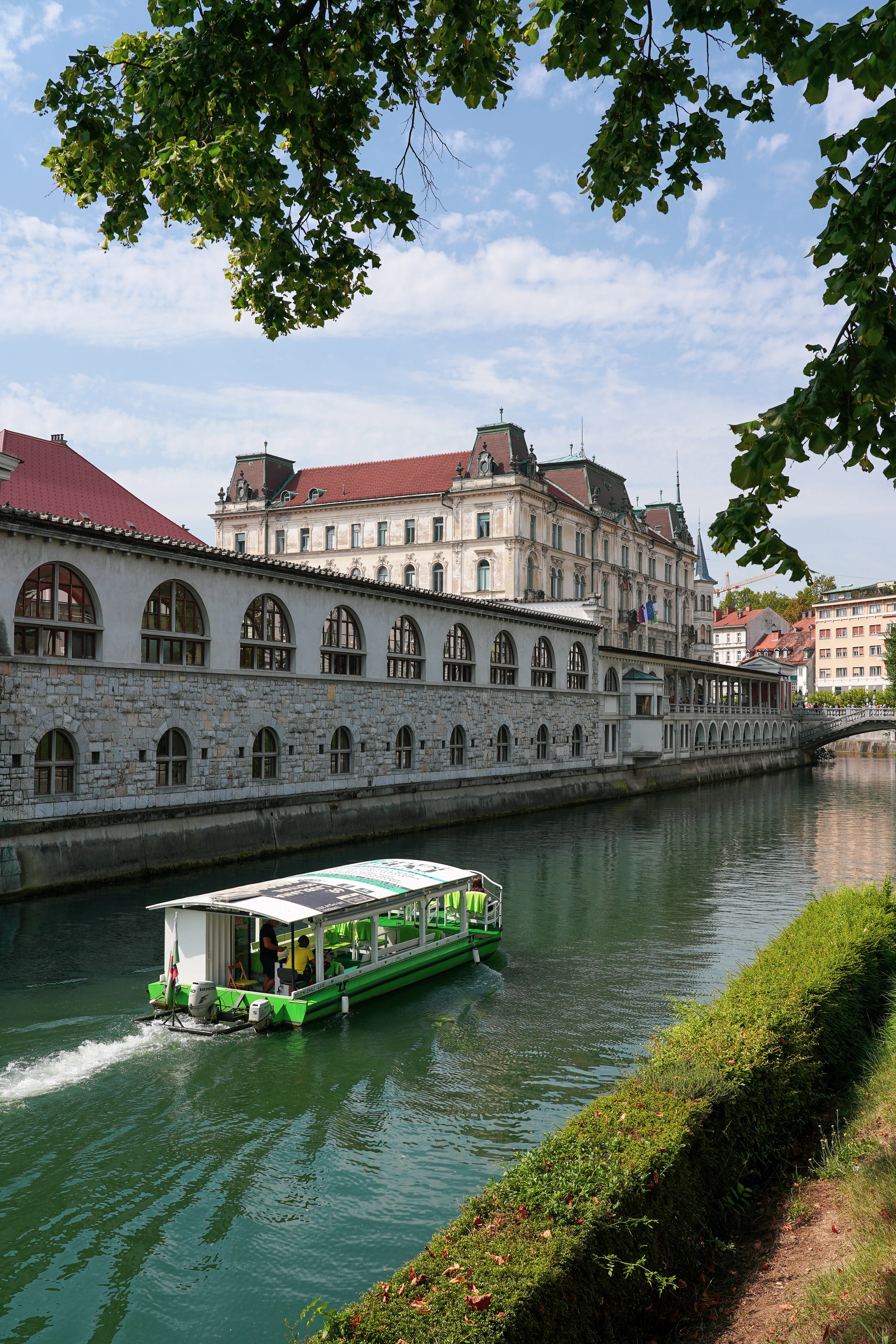 A tour boat gliding along the green Ljubljanica river past the arcades of the riverside Central Market in Ljubljana