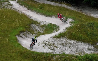 Two mountain bikers navigate a winding dirt trail surrounded by green grass and sparse vegetation. The path takes several sharp turns, providing a dynamic scene. The bikers wear helmets and sport attire, adding a sense of motion and adventure.
