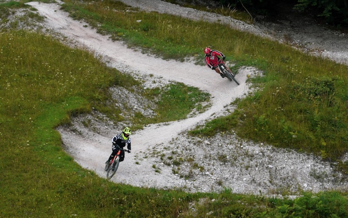 Two mountain bikers navigate a winding dirt trail surrounded by green grass and sparse vegetation. The path takes several sharp turns, providing a dynamic scene. The bikers wear helmets and sport attire, adding a sense of motion and adventure.