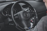 A detailed view of a car's interior focusing on the steering wheel, which has the Opel logo. Adjacent to the steering wheel, various control switches and a glimpse of the dashboard with dials and gauges are visible. Part of a person's shoulder is also in view.
