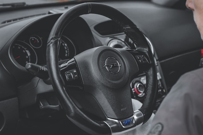 A detailed view of a car's interior focusing on the steering wheel, which has the Opel logo. Adjacent to the steering wheel, various control switches and a glimpse of the dashboard with dials and gauges are visible. Part of a person's shoulder is also in view.