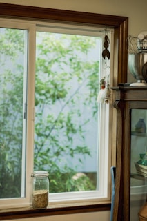 A bright and cozy kitchen scene featuring a window with a view of green foliage outside. The window is framed with brown wood, and a glass jar filled with seeds or grains sits on the windowsill. To the right, part of a wooden cabinet is visible, holding kitchen items. A decorative hanging with feathers and small elements adds an artistic touch above the jar.