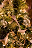 A lush, heavily decorated holiday tree featuring green foliage, white floral elements, and warm white string lights. The tree has elegant beige ribbons woven through the branches, complemented by geometric gold ornaments. The overall theme is natural and elegant, with an emphasis on greenery and warm lighting.