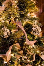 A lush, heavily decorated holiday tree featuring green foliage, white floral elements, and warm white string lights. The tree has elegant beige ribbons woven through the branches, complemented by geometric gold ornaments. The overall theme is natural and elegant, with an emphasis on greenery and warm lighting.