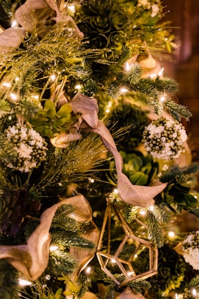 A lush, heavily decorated holiday tree featuring green foliage, white floral elements, and warm white string lights. The tree has elegant beige ribbons woven through the branches, complemented by geometric gold ornaments. The overall theme is natural and elegant, with an emphasis on greenery and warm lighting.