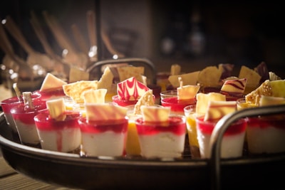 A tray filled with elegantly arranged small dessert cups featuring layers of white and red mousse, garnished with intricately designed chocolate pieces and wafers. The desserts are creatively presented and illuminated under soft, warm lighting, highlighting their vibrant colors and textures.