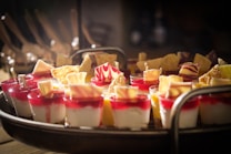 A tray filled with elegantly arranged small dessert cups featuring layers of white and red mousse, garnished with intricately designed chocolate pieces and wafers. The desserts are creatively presented and illuminated under soft, warm lighting, highlighting their vibrant colors and textures.