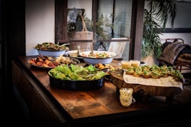 A wooden table is elegantly arranged with various dishes, including salads in bowls and appetizers on a rustic wooden platter. There are leafy greens, roasted vegetables, and what appears to be puddings or dessert cups. The setting has a cozy, rustic atmosphere with natural light coming through a window, plants in the background, and a coat draped over a chair.