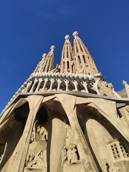 A detailed view of the Sagrada Familia's intricate architecture with towering spires reaching into a clear blue sky. The lower portion features ornate carvings and structural columns.
