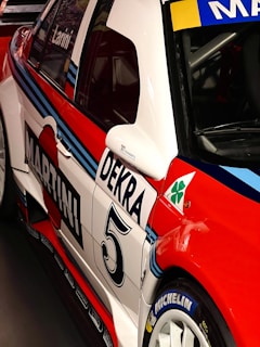 A close-up view of a racing car adorned with vibrant red, white, and blue colors. The car features prominent branding such as 'Martini' and 'Dekra' along with various sponsorship logos. The number '5' is clearly visible, and the car’s body is sleek and aerodynamic, designed for high performance.