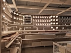 A well-organized display of various lighting fixtures, including wall-mounted lamps, spotlights, and ceiling lights, arranged on a dark wall. Shelves beneath the lights hold boxed products, and a few price tags are visible. The setting appears to be a store showroom with a focus on lighting solutions.