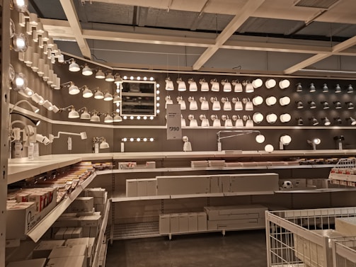 A well-organized display of various lighting fixtures, including wall-mounted lamps, spotlights, and ceiling lights, arranged on a dark wall. Shelves beneath the lights hold boxed products, and a few price tags are visible. The setting appears to be a store showroom with a focus on lighting solutions.