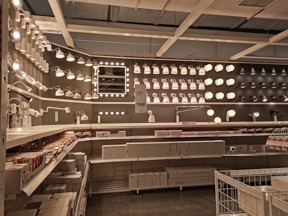 A well-organized display of various lighting fixtures, including wall-mounted lamps, spotlights, and ceiling lights, arranged on a dark wall. Shelves beneath the lights hold boxed products, and a few price tags are visible. The setting appears to be a store showroom with a focus on lighting solutions.