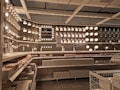A well-organized display of various lighting fixtures, including wall-mounted lamps, spotlights, and ceiling lights, arranged on a dark wall. Shelves beneath the lights hold boxed products, and a few price tags are visible. The setting appears to be a store showroom with a focus on lighting solutions.