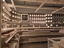A well-organized display of various lighting fixtures, including wall-mounted lamps, spotlights, and ceiling lights, arranged on a dark wall. Shelves beneath the lights hold boxed products, and a few price tags are visible. The setting appears to be a store showroom with a focus on lighting solutions.