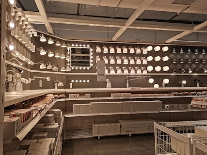 A well-organized display of various lighting fixtures, including wall-mounted lamps, spotlights, and ceiling lights, arranged on a dark wall. Shelves beneath the lights hold boxed products, and a few price tags are visible. The setting appears to be a store showroom with a focus on lighting solutions.