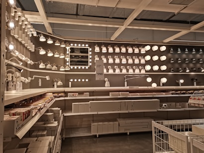 A well-organized display of various lighting fixtures, including wall-mounted lamps, spotlights, and ceiling lights, arranged on a dark wall. Shelves beneath the lights hold boxed products, and a few price tags are visible. The setting appears to be a store showroom with a focus on lighting solutions.