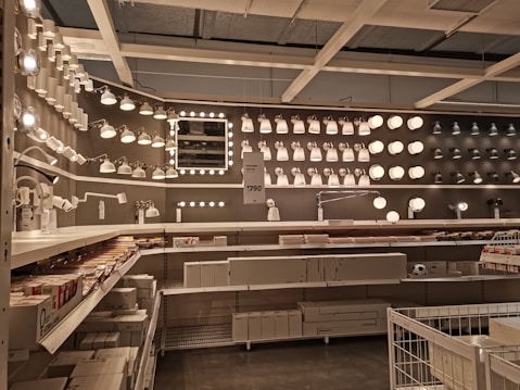 A well-organized display of various lighting fixtures, including wall-mounted lamps, spotlights, and ceiling lights, arranged on a dark wall. Shelves beneath the lights hold boxed products, and a few price tags are visible. The setting appears to be a store showroom with a focus on lighting solutions.