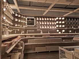 A well-organized display of various lighting fixtures, including wall-mounted lamps, spotlights, and ceiling lights, arranged on a dark wall. Shelves beneath the lights hold boxed products, and a few price tags are visible. The setting appears to be a store showroom with a focus on lighting solutions.