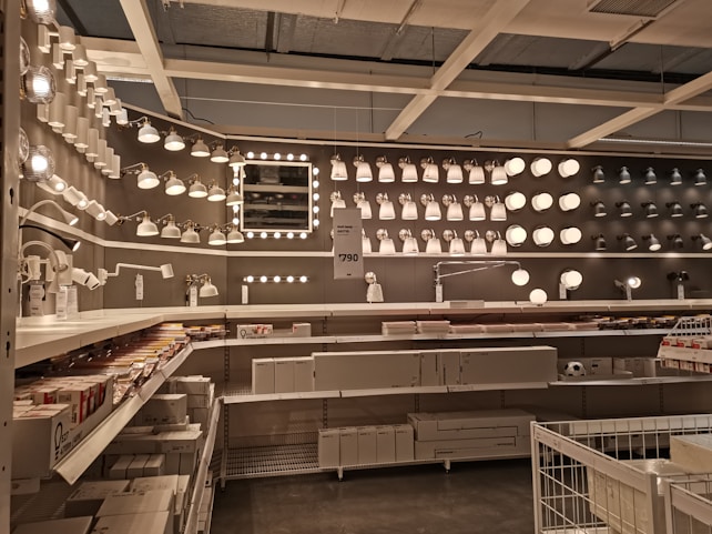 A well-organized display of various lighting fixtures, including wall-mounted lamps, spotlights, and ceiling lights, arranged on a dark wall. Shelves beneath the lights hold boxed products, and a few price tags are visible. The setting appears to be a store showroom with a focus on lighting solutions.