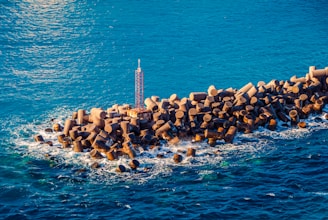 A breakwater structure made of evenly arranged large cylindrical concrete blocks extends into the vibrant blue sea, with waves crashing against the blocks. The structure includes a tall, narrow metal tower at the center.