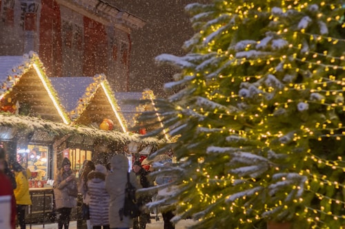 Snow falls gently over a festive outdoor market, with small wooden stalls decorated with bright, warm lights. People are dressed in winter clothing, enjoying the atmosphere. A large Christmas tree dominates the foreground, adorned with numerous yellow lights. The scene evokes a quintessential holiday ambiance.